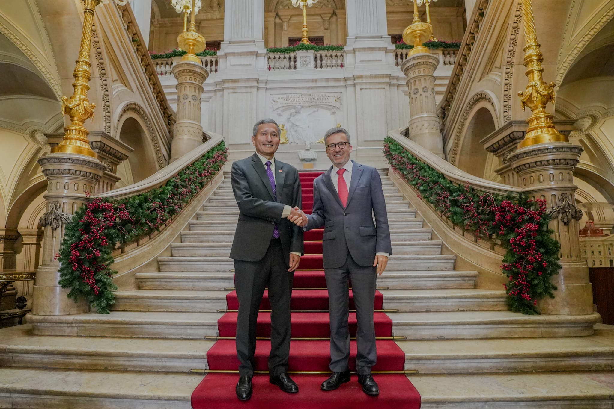 Two men in suits shake hands on red-carpeted stairs decorated with greenery.